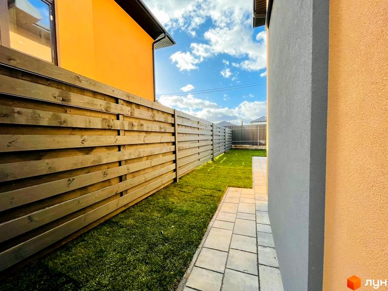 Дуплекси „Карамель Сіті“: зовнішній вигляд будинку з оранжевим фасадом та дерев'яним парканом, доглянута галявина та бруківка.