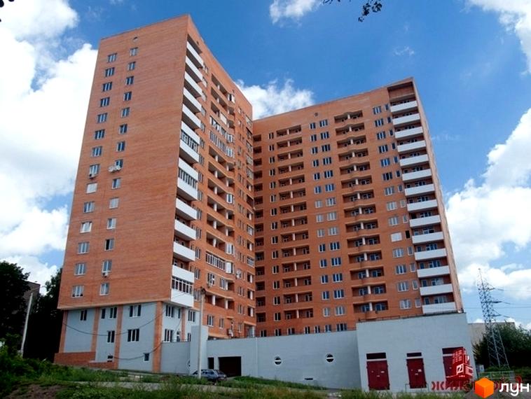 Новобудова Гранд: багатоповерховий будинок L-подібної форми з цегляним фасадом, білими балконами та вікнами. Навколо зелена територія та господарські будівлі.