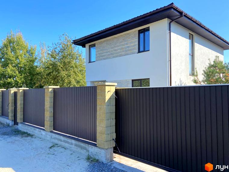 Вигляд двоповерхового будинку з коричневим парканом та колонами з цегли. На задньому плані зелені дерева та блакитне небо.