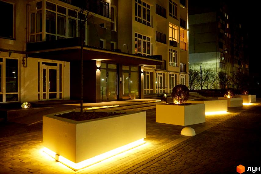 Житловий комплекс KromaxBud. Зовнішній вигляд новобудови вночі з підсвіченими клумбами та входом. Територія комплексу освітлена.