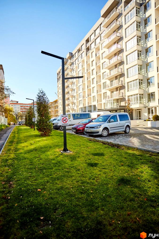 Зображення фасаду та прибудинкової території Житлового комплексу KromaxBud. Багатоповерховий будинок з балконами, поруч зелений газон, дерева та припарковані авто.