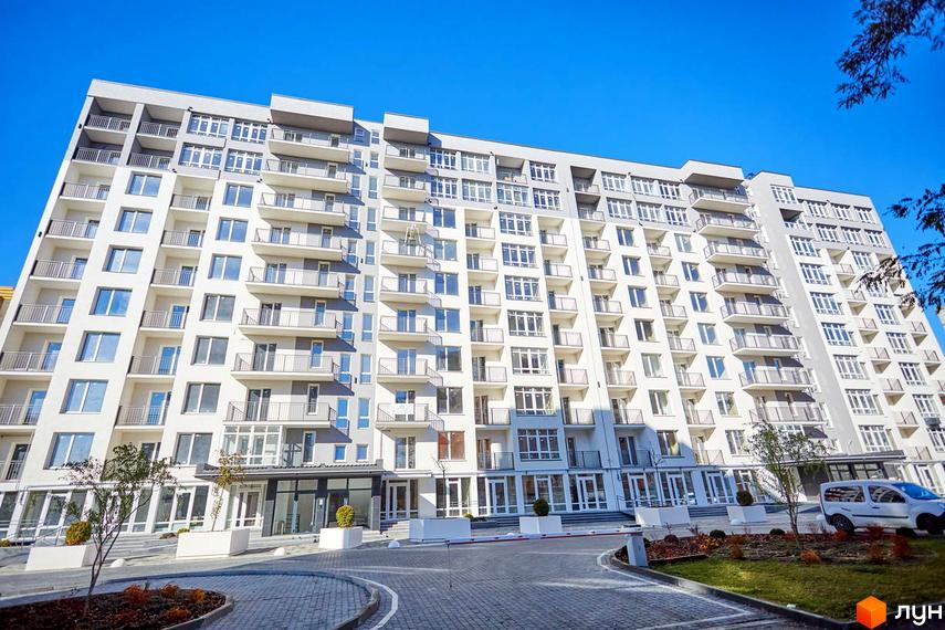 Новобудова ЖК KromaxBud з численними балконами та великими вікнами під блакитним небом. Перед будинком облаштована прибудинкова територія з зеленими насадженнями та доріжками.