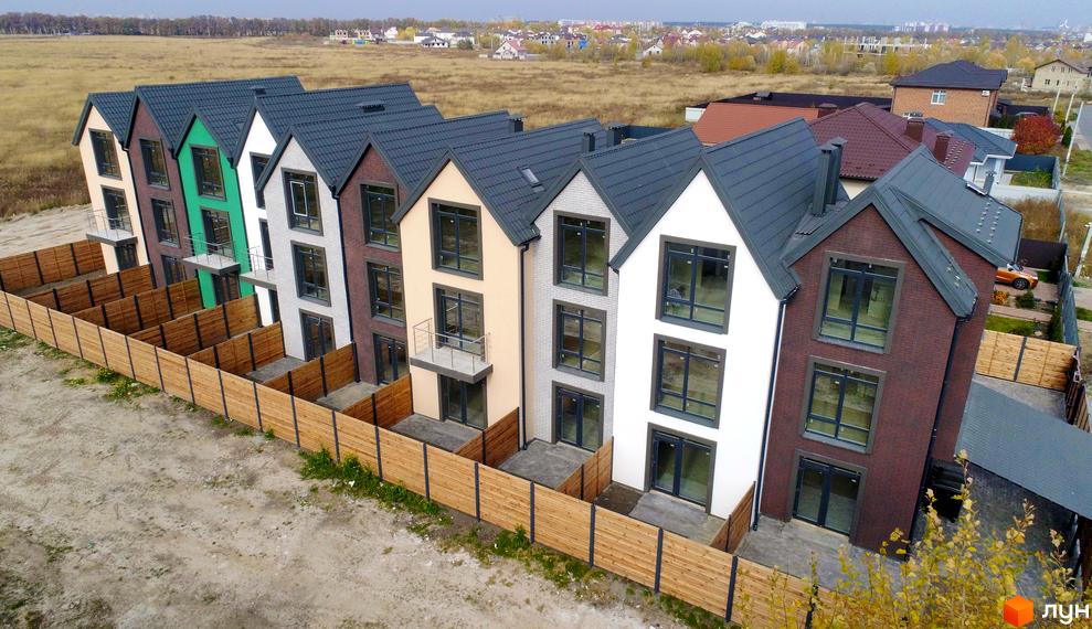 Зображення таунхаусів New Smart X з різнокольоровими фасадами та темними дахами. Перед будинками розташовані невеликі дворики, огороджені дерев'яним парканом.