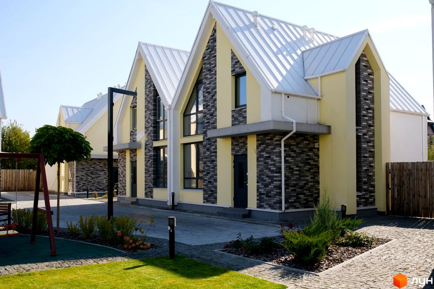 Будинки Котеджного містечка Solo Home мають світло-жовті фасади з сірою цеглою та білі дахи. На прибудинковій території видно зелені газони, кущі та дитячий майданчик.