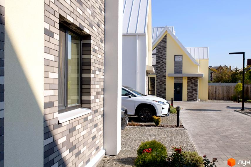 Котеджне містечко Solo Home: будинок з жовтими стінами та цегляним фасадом сіро-білих відтінків, біля якого припаркований автомобіль на вимощеній бруківкою території.