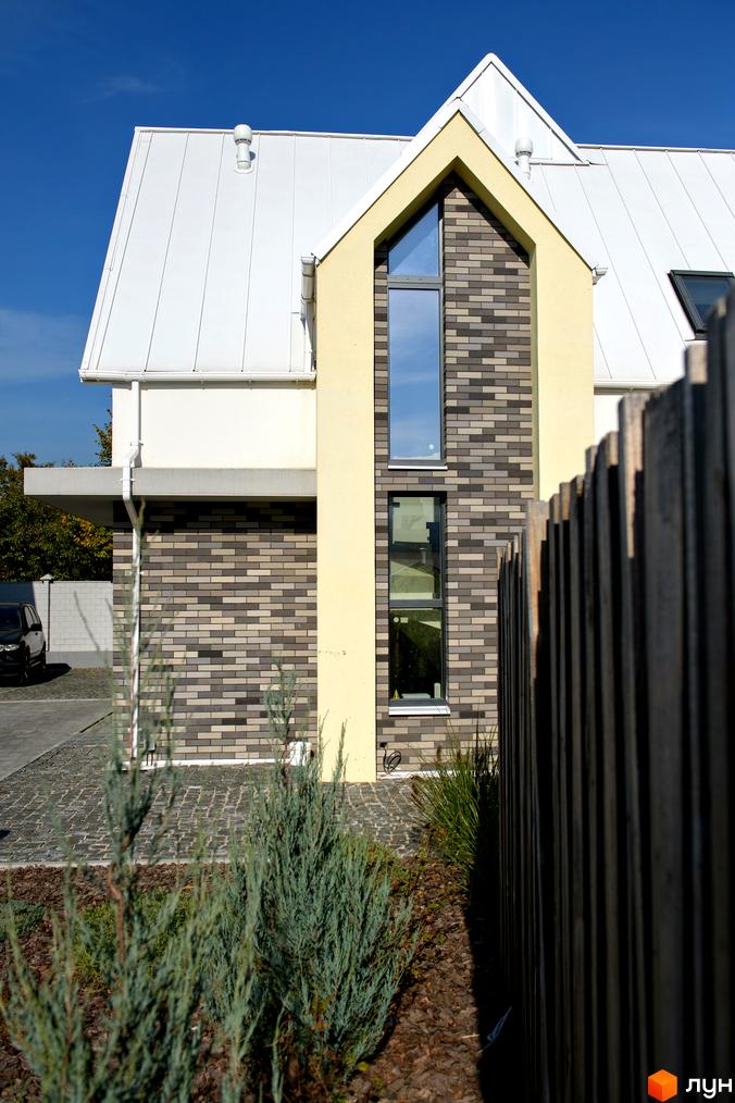 Будинок Котеджного містечка Solo Home з білим дахом та фасадом з жовтої штукатурки і сірої цегли. Перед будинком розташовані зелені насадження та дерев'яний паркан.