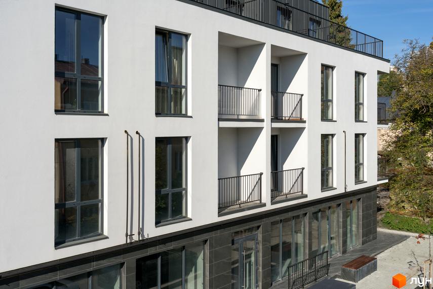 Житловий комплекс Globus Elite має білий фасад з великими вікнами та балконами з чорними поручнями. Прибудинкова територія облаштована лавкою та зеленню.