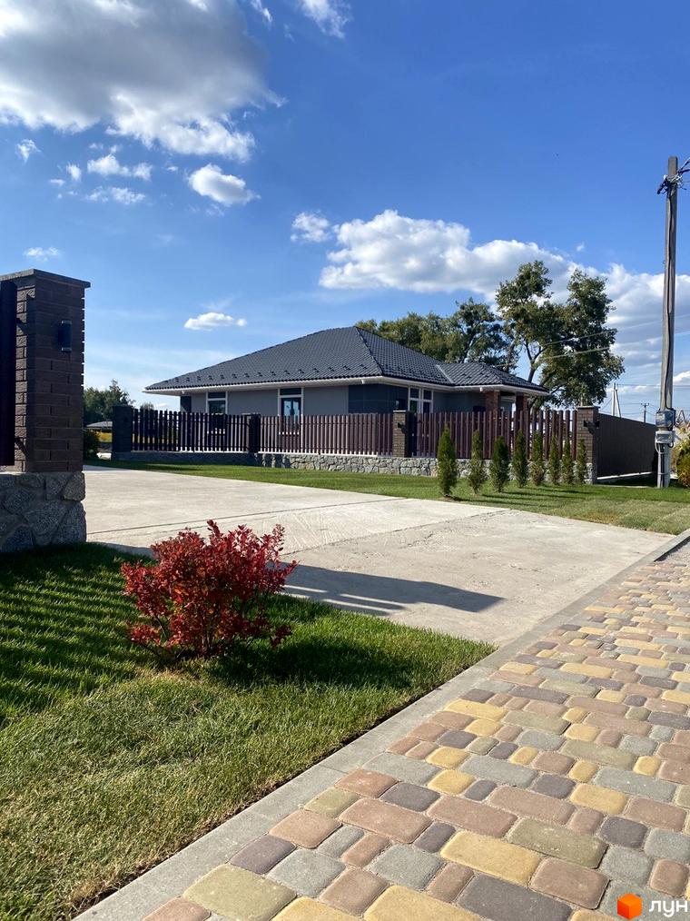 Одноповерховий будинок з темним дахом та парканом, доглянута прибудинкова територія з газоном та бруківкою. Містечко котеджів Крушинка Парк.