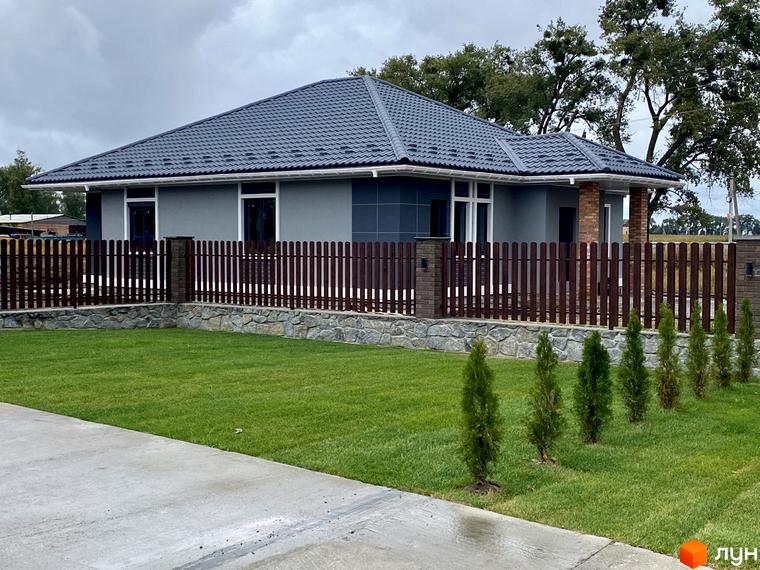 Одноповерховий будинок з темною черепицею та сірими стінами. Перед будинком зелена галявина з невеликими туями та коричневий паркан з кам'яним фундаментом.