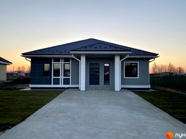 Одноповерховий будинок з сірим фасадом та темною черепицею в Котеджному комплексі Крушинка Парк. Перед будинком бетонна доріжка та зелений газон.