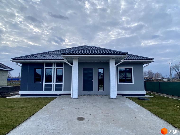 Одноповерховий будинок з темною покрівлею та сірими стінами. Перед будинком бетонна доріжка, газон та хмарне небо.