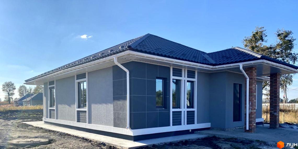 Одноповерховий будинок з темно-сірою черепичною покрівлею та світлими стінами. На прибудинковій території видно землю та траву.