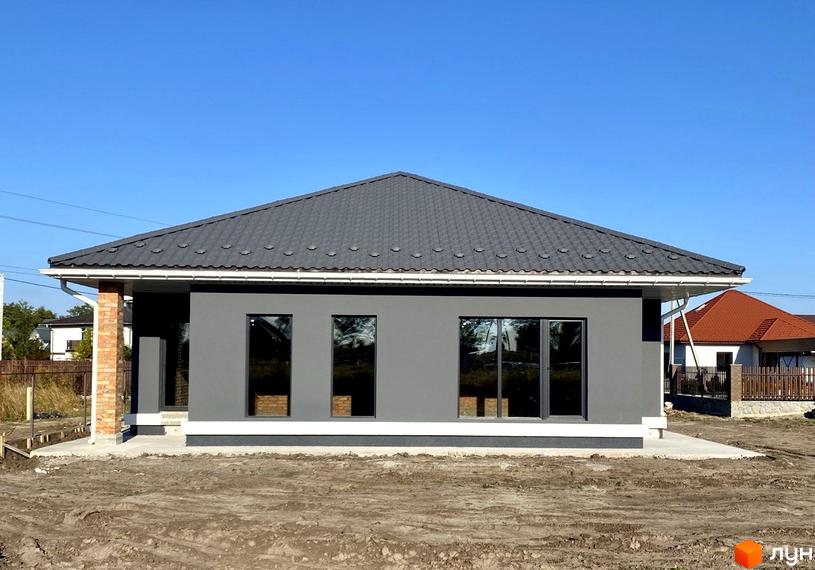 Одноповерховий будинок з сірими стінами та темним дахом. Перед будинком розташована ділянка землі, на задньому плані видно інші будинки.