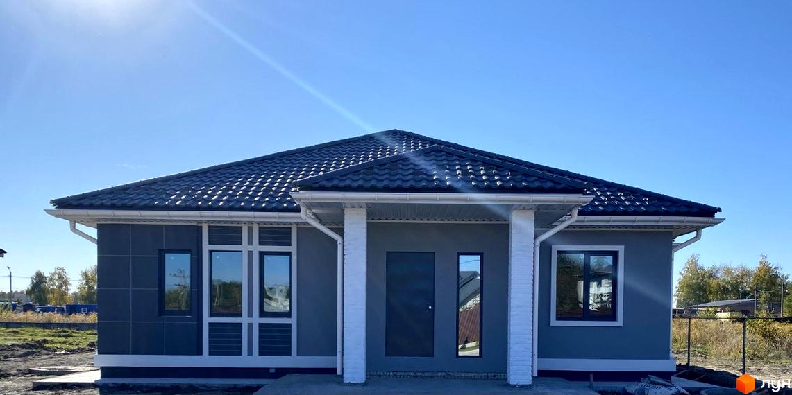 Одноповерховий будинок з темною покрівлею та сірими стінами. Біля будинку газон та дерева, сонячна погода.