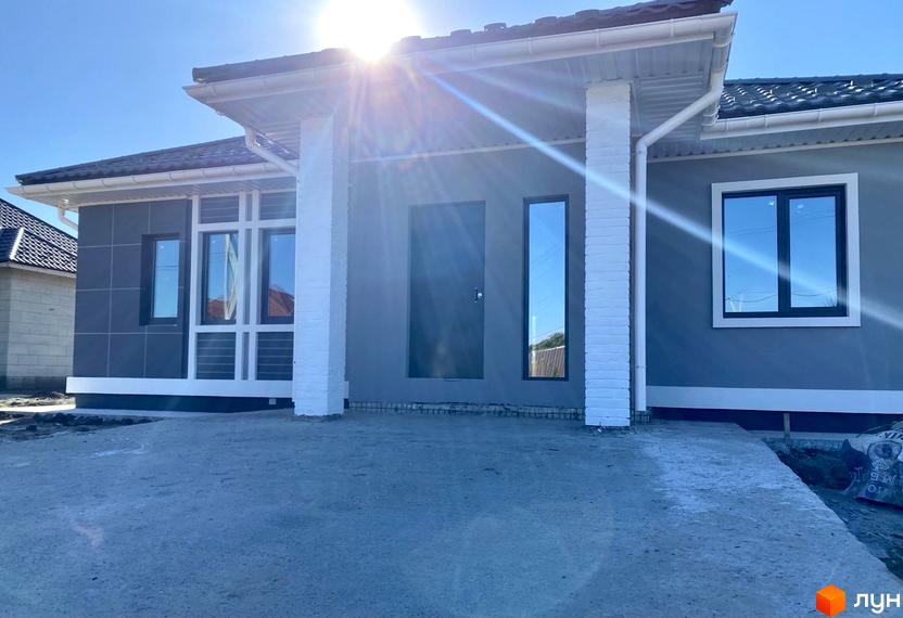 Фасад будинку в Котеджному містечку Крушинка Парк з сірими стінами, білими колонами та вікнами з темними рамами, перед будинком бетонна площадка.