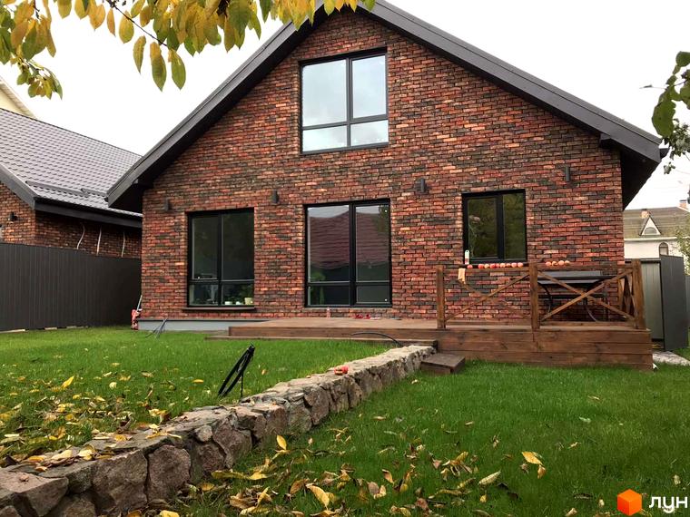 Зовнішній вигляд будинку з червоної цегли з великими вікнами та дерев'яною терасою. Навколо будинку зелений газон з опалим листям та кам'яною огорожею.