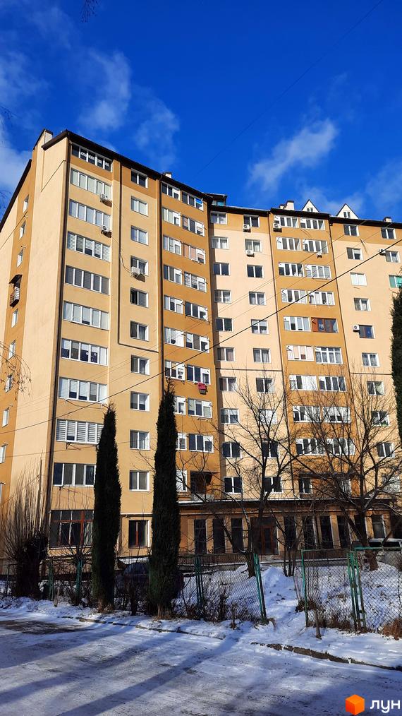 Зовнішній вигляд багатоповерхового будинку на вулиці Чорновола, 128а, з вікнами та кондиціонерами. Прибудинкова територія вкрита снігом, з деревами та парканом.