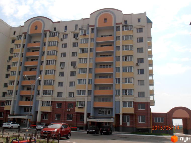 Житловий комплекс Юність з фасадом у світлих тонах та великими вікнами. Прибудинкова територія з автомобілями та освітленням.