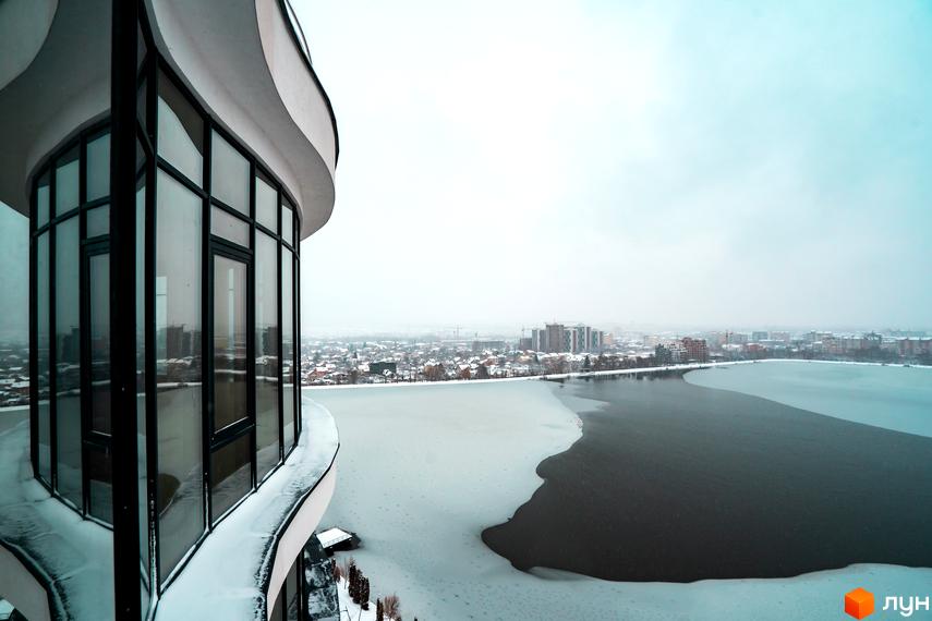 Зображення житлового комплексу Parus взимку. Вид на новобудову з великими вікнами та засніжене озеро.