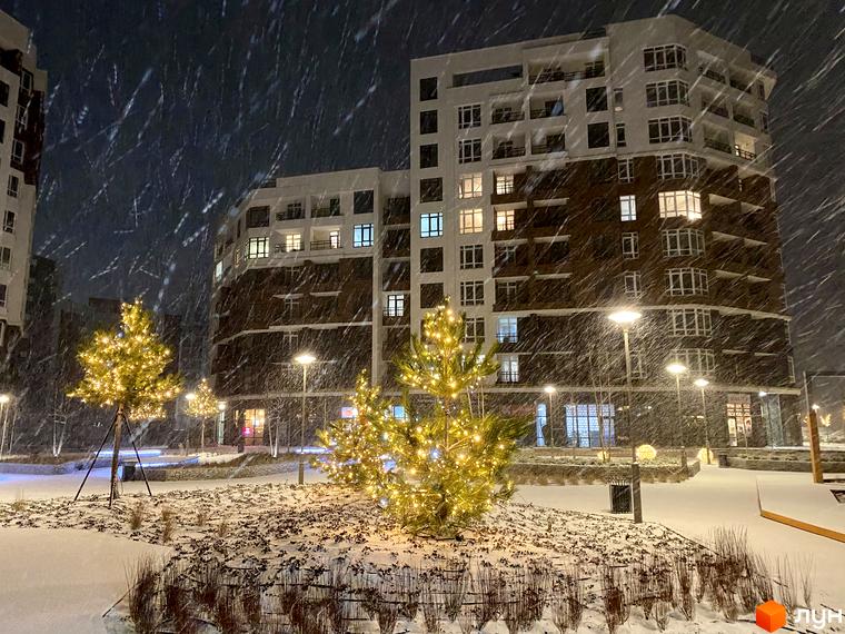Житловий комплекс Синергія Сіті взимку під час снігопаду. Навколо новобудови прикрашені ялинки з гірляндами та освітлення.