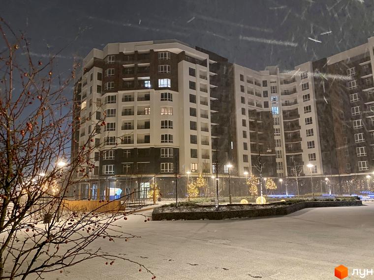 Фасад житлового комплексу Синергія Сіті взимку під час снігопаду. Навколо будівлі розташована прибудинкова територія з деревами та освітленням.