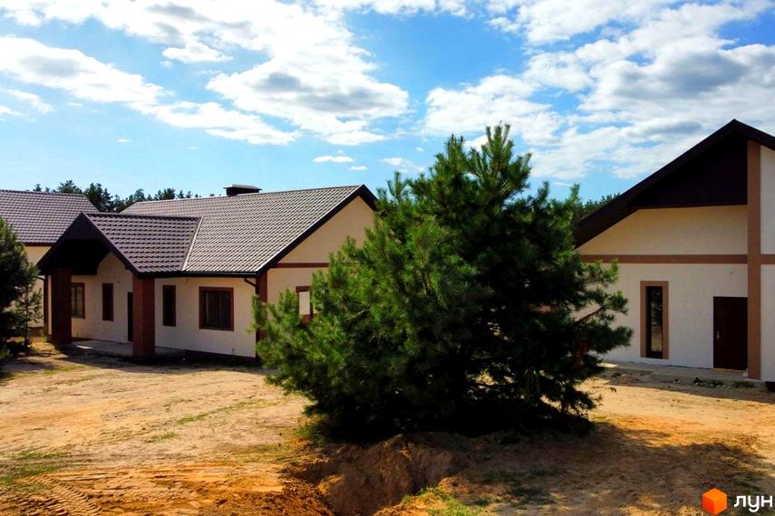 Котеджне містечко КМ Лісовий Масив: двоповерхові будинки з коричневими дахами на фоні блакитного неба з хмарами. Навколо будівельний майданчик з піщаним покриттям та зелені дерева.