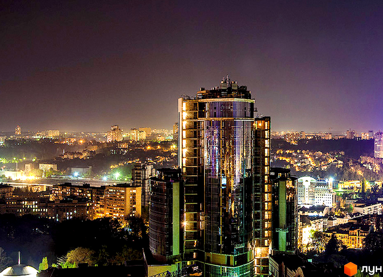 Нічний вид на висотну будівлю Житловий комплекс H-Tower з дзеркальним фасадом та освітленими вікнами. Навколо видно міські забудови та зелені насадження.