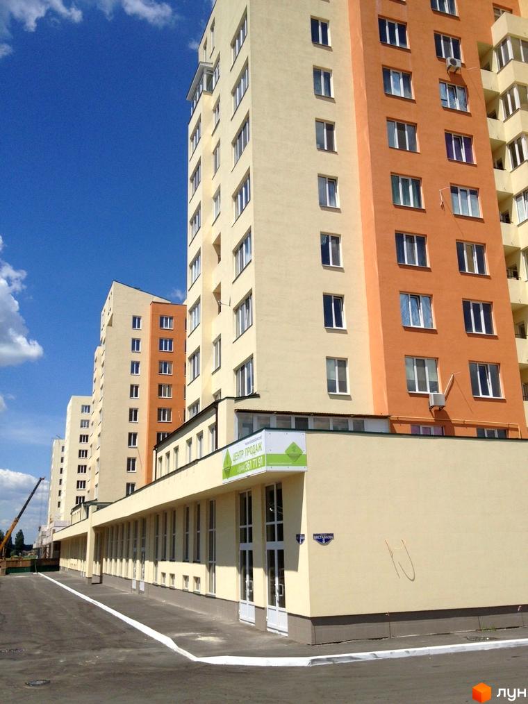 Житловий комплекс Новосельцево з фасадом жовтого та оранжевого кольору. Прибудинкова територія з асфальтованою доріжкою та вікнами на першому поверсі.