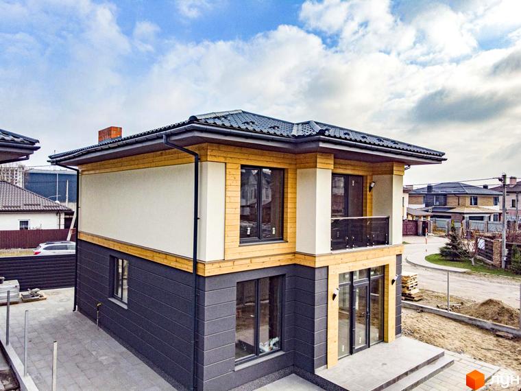 Котеджне містечко КМ Family Villas: двоповерховий будинок з дерев'яними та сірими фасадами, великими вікнами та балконом. Навколо будинку – бруківка та зелені насадження.