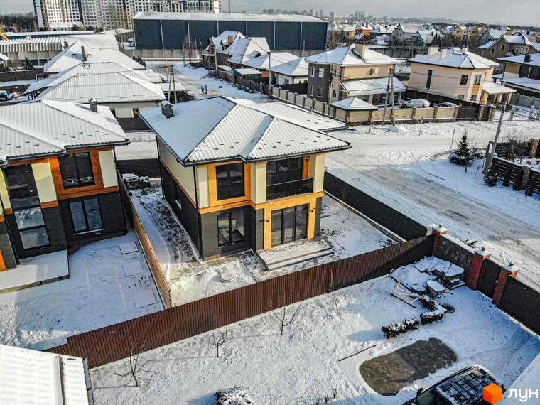 Котеджне містечко КМ Family Villas у зимовий період. Двоповерховий будинок з темним та світлим оздобленням фасаду, великими вікнами та балконом, на тлі засніженої прибудинкової території та вулиці.