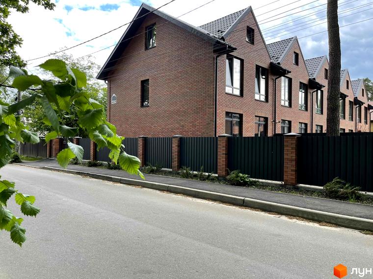 Фасад таунхаусів BRIGHTON GARDEN: цегляні будинки з темними дахами та вікнами. Перед ними зелений паркан, тротуар і дорога.