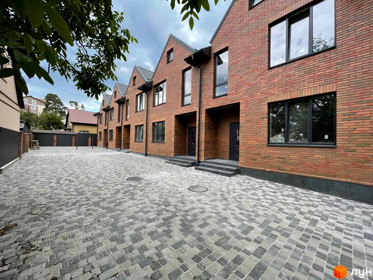 Ряд цегляних таунхаусів Таунхауси ELEGANT з темними дахами та вікнами, перед якими вимощена бруківкою прибудинкова територія.