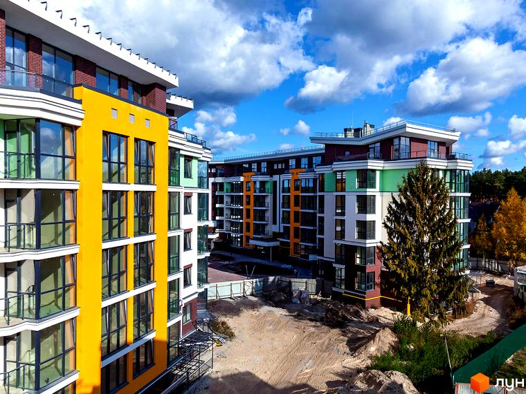 Житловий комплекс Лісова казка 2 - сучасна новобудова з яскравим фасадом та великими вікнами. Навколо будинку - будівельний майданчик з деревами та хмарним небом.