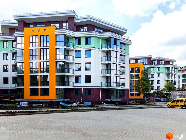 Житловий комплекс Лісова казка 2. Фасад будинку з яскравими елементами, панорамними вікнами та балконами. Прибудинкова територія з бруківкою та зеленими насадженнями.