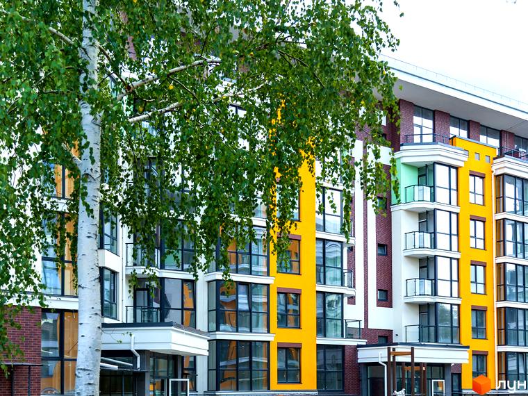 Фасад житлового комплексу Лісова казка 2 з яскравими жовтими та білими елементами, великими вікнами та балконами. Зелене листя дерев прикрашає будівлю.