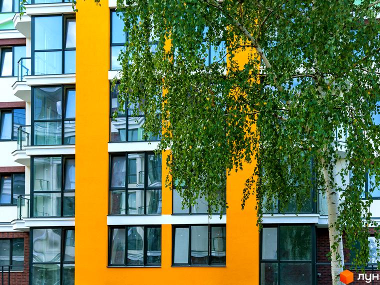 Фасад новобудови Лісова казка 2 з яскраво-жовтою вертикальною вставкою та великими вікнами. На передньому плані гілки дерева з зеленим листям.