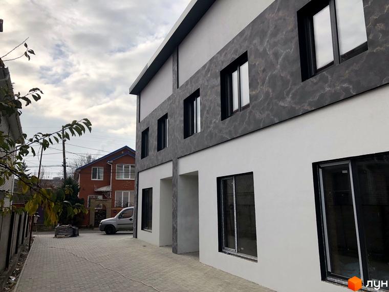 Зовнішній вигляд новобудови на вул. Підлісна (Анастасії Медвідь), 32, з сірим фасадом та білими стінами. Поруч розташована брукована доріжка та червоний будинок.