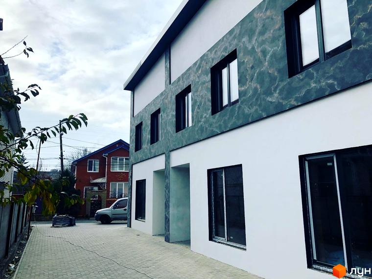 Зовнішній вигляд новобудови на вул. Підлісна (Анастасії Медвідь), 32, з сірою оздобленою фасадною панеллю та білими стінами. Поруч розташована брукована доріжка та червоний двоповерховий будинок.