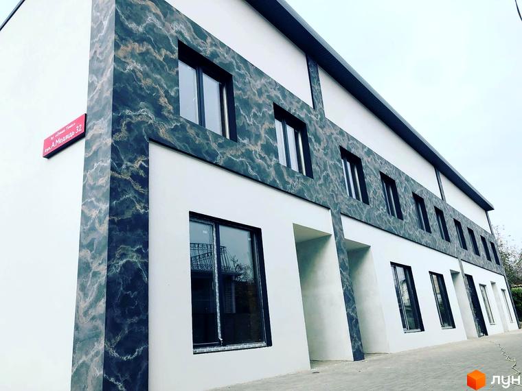 Фасад житлового комплексу на вул. Анастасії Медвідь, 32, з білими стінами та темними панелями під мармур. Перед будинком вимощена територія.