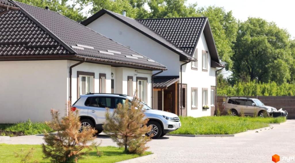 Котеджне містечко КМ Титул: фасади білих будинків з темними дахами та прибудинкова територія з автомобілями та зеленими насадженнями.