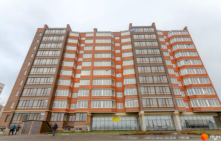 Зовнішній вигляд багатоповерхового будинку ЖК Петрівський Квартал з червоної цегли з великою кількістю вікон. Поруч з будинком видно вхід до під'їздів та людей, що йдуть.