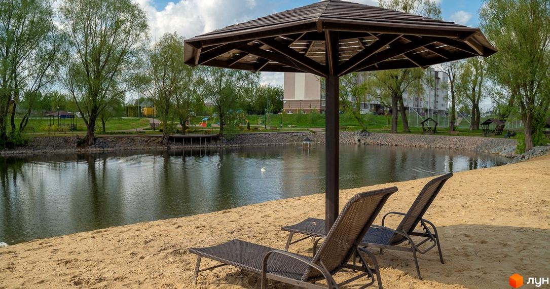 Житловий комплекс Озерний Гай (Гатне) з багатоповерховими будинками. Перед ними піщаний пляж, озеро та зелена зона відпочинку з деревами.