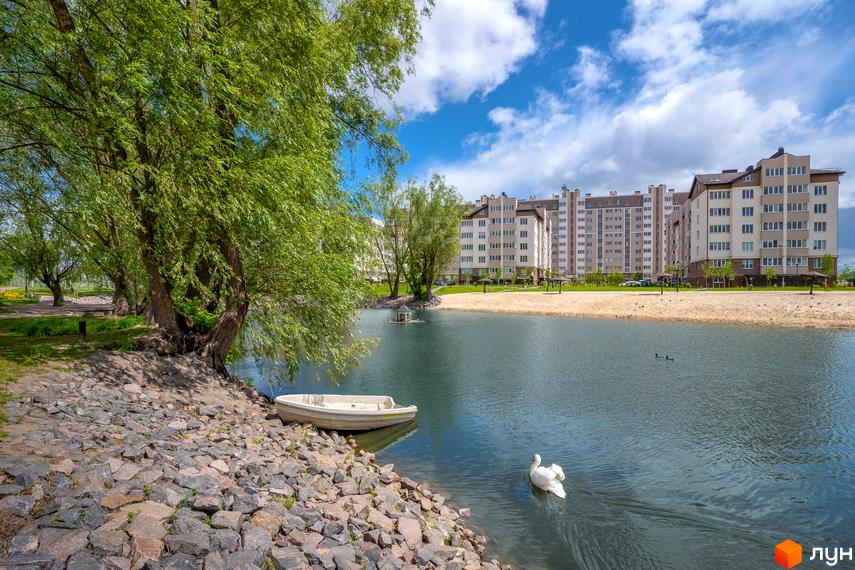 Багатоповерхові будинки Житлового комплексу Озерний Гай (Гатне) на березі озера. Навколо зелені дерева, піщаний пляж, човен та лебідь на воді.