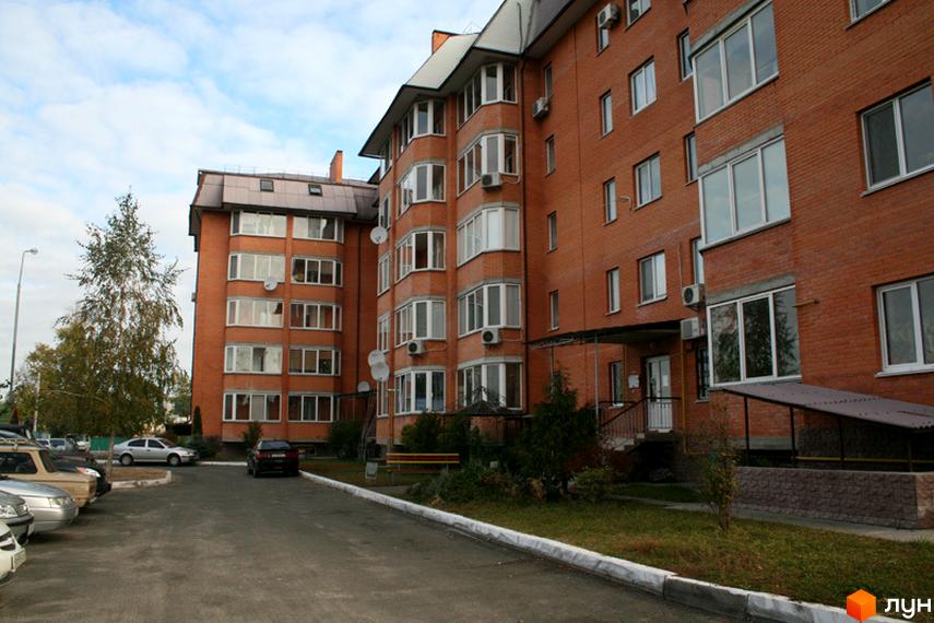 Багатоповерховий цегляний житловий будинок з балконами та вікнами. Перед будинком є асфальтований проїзд з припаркованими автомобілями та зелена зона з деревами.