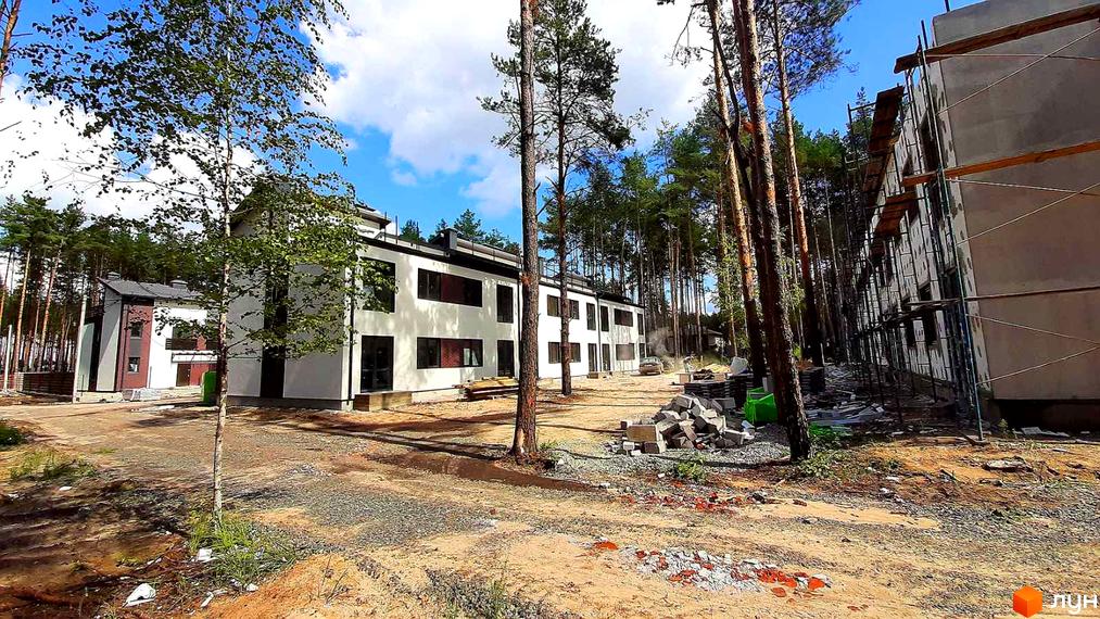 Зображення сторінки житлового комплексу КМ Лісове-2. Білі будинки з темними вікнами розташовані серед дерев. На території видно будівельні матеріали та пісок.