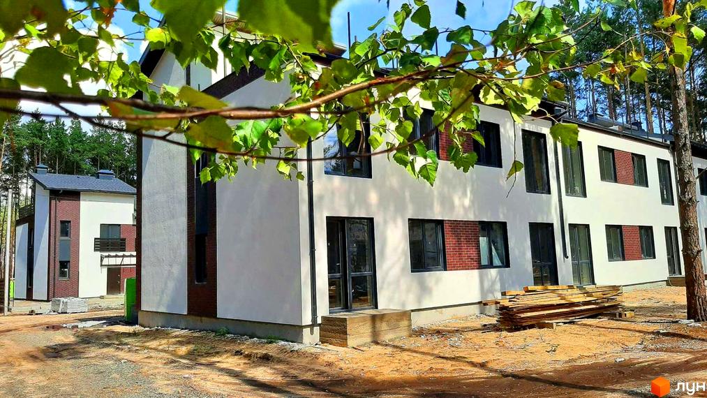 Зовнішній вигляд будинку в Котеджному містечку Лісове-2. Двоповерховий будинок з білими стінами та чорними вікнами, поруч дерева та будівельні матеріали.