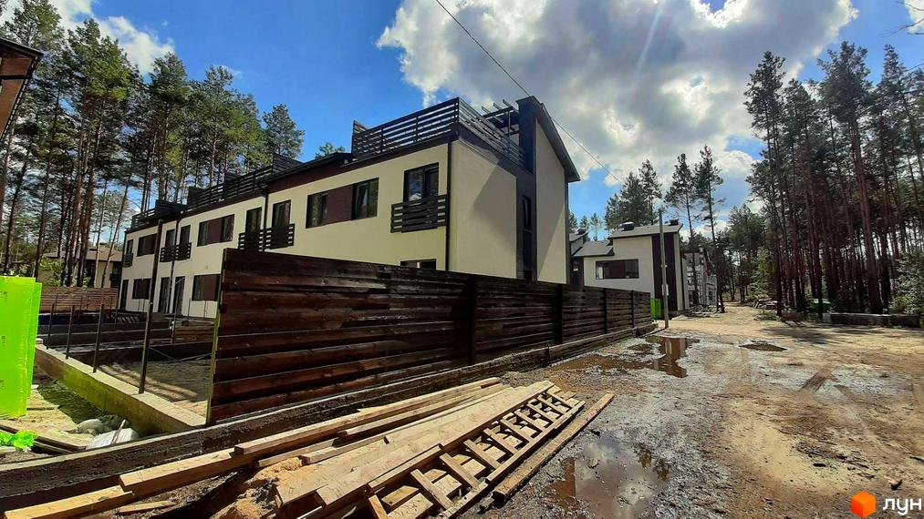 Зображення сторінки житлового комплексу КМ Лісове-2. Будинки з темним дахом та дерев'яним парканом. На території видно будівельні матеріали та дерева.