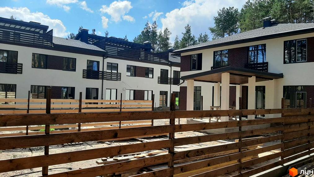 Зображення житлового комплексу з білими стінами та коричневим дахом. Перед будинком дерев'яний паркан, а на задньому плані видно дерева та небо з хмарами.