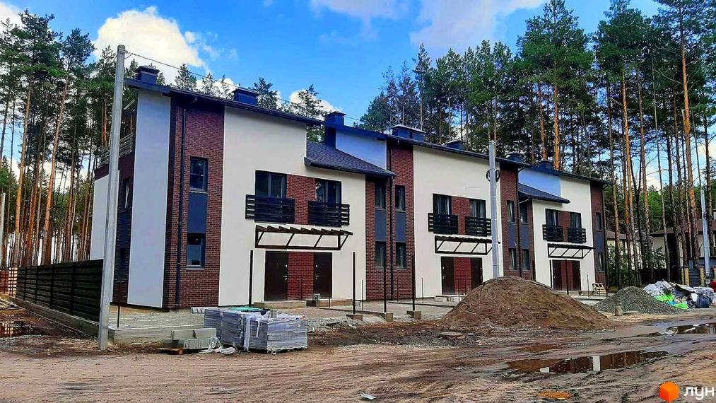 Зображення Котеджне містечко Лісове-2: триповерхові таунхауси з червоної цегли та білою штукатуркою, чорні металеві балкони та навіси над вхідними дверима. Навколо сосновий ліс та будівельні матеріали.