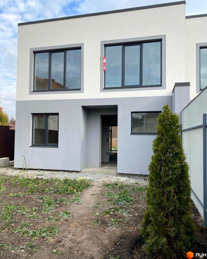 Котеджне містечко Чапаєвка: двоповерховий будинок з сірими вікнами та сірою частиною фасаду, на тлі блакитного неба. Перед будинком зелена ялина та ділянка з рослинністю.